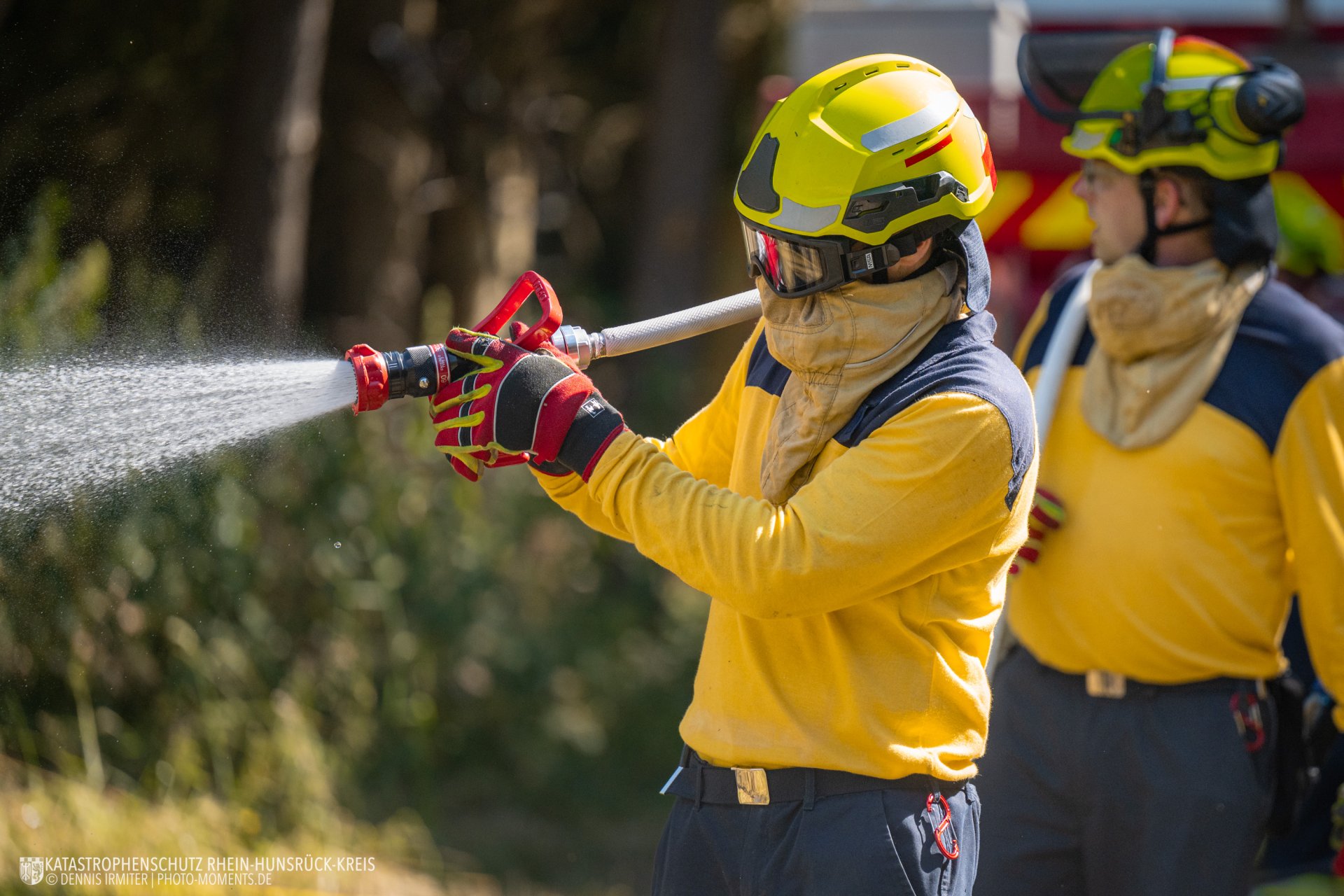 Eine Einsatzkraft in gelber Schutzkleidung, mit Helm, Schutzbrille und Atemschutz, hält einen Feuerwehrschlauch bei einem Löscheinsatz im Freien.
