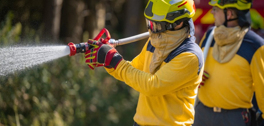 Waldbrandübung Eine Einsatzkraft in gelber Schutzkleidung, mit Helm, Schutzbrille und Atemschutz, hält einen Feuerwehrschlauch bei einem Löscheinsatz im Freien.